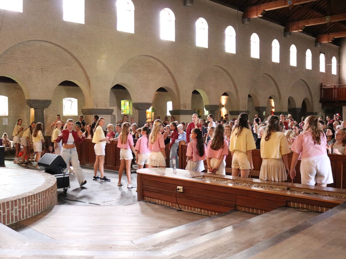 Een volle kerk tijdens de dansvoorstelling: dansers in roze en geel op de vloer, publiek in de banken.